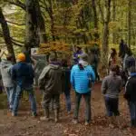 Un groupe de gens écoute la présentation d'un homme devant un tableau, en bordure de forêt.