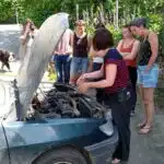 7 femmes sont regroupées dehors autour d'une voiture. Le capot de cette voiture est ouvert et une femme est penchée dessus, en train d'expliquer quelque chose aux autres femmes.